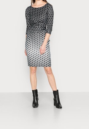 Woman wearing a black and white geometric patterned knee-length dress with three-quarter sleeves and black ankle boots standing on a plain background.
