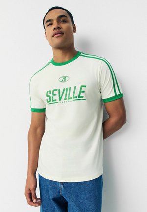Biela tričko s zelenými akcentmi, s potlačou "SEVILLE" a "79" na prednej strane. Krátke rukávy a okrúhly výstrih.