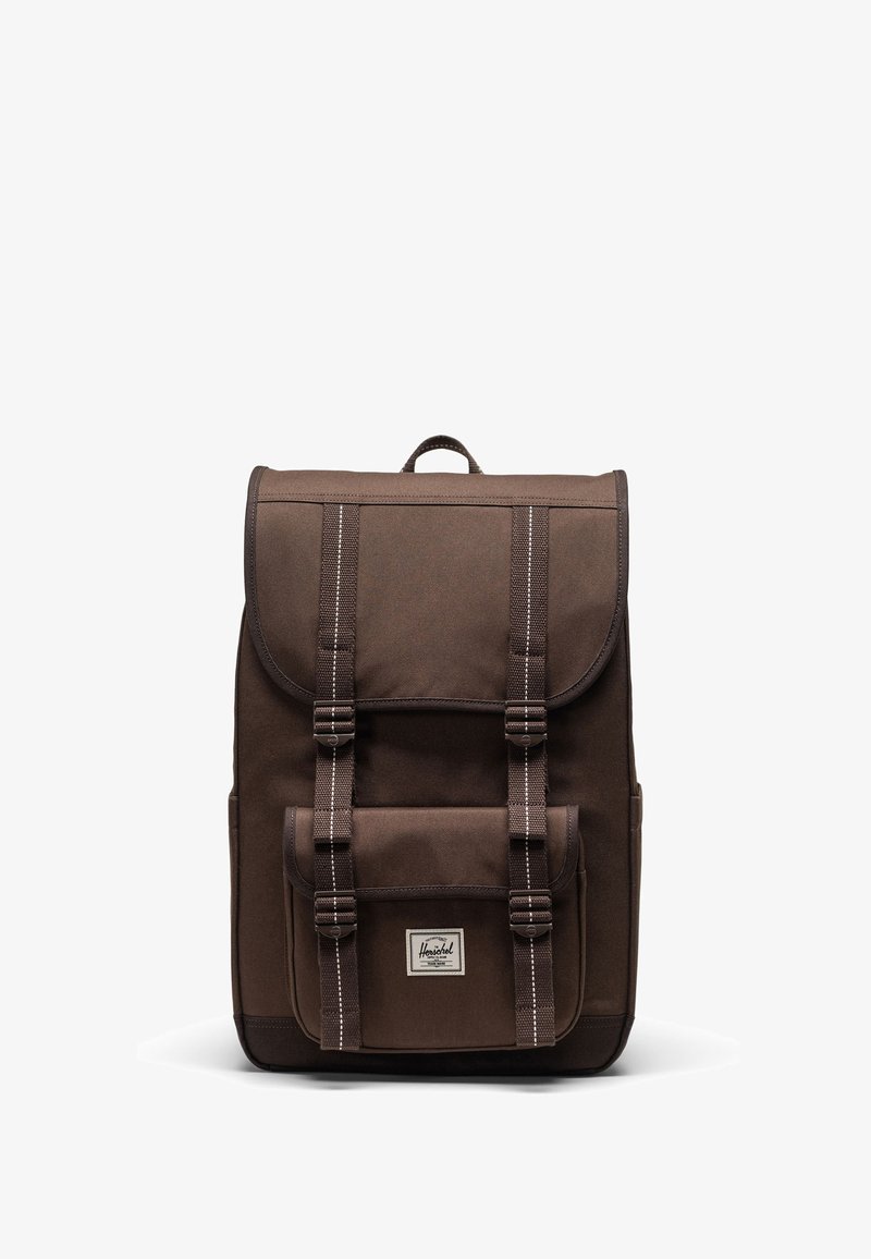 Brun canvasryggsäck med rulltoppdesign, som har två fram- och spännband, en framficka och en tygetikett vid basen.