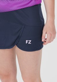 Laivastonsininen urheiluhame, jossa on aaltoileva helma. Hameessa on hienovarainen tekstuurikuosi ja vaaleanpunainen "FZ" -logo vasemmassa alareunassa.