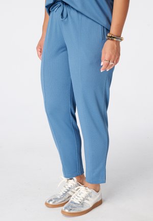 Persoon die blauwe gestructureerde broek draagt met een trekkoord in de taille, zilveren en witte sneakers en armbanden om de pols.