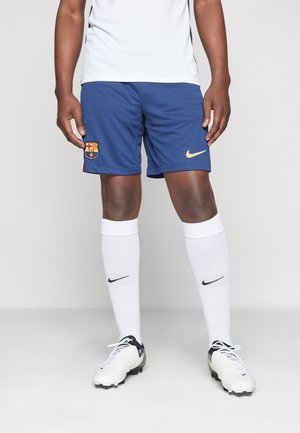 Mężczyzna w białej sportowej koszulce, niebieskich spodenkach FC Barcelona z logo Nike, białych podkolanówkach Nike i białych korkach stoi na szarej podłodze.