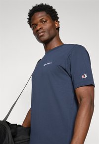 Marineblaue Baumwoll-T-Shirt mit kurzen Ärmeln und Rundhalsausschnitt, versehen mit weißem, geschwungenem "Champion"-Logo auf der Brust und Logo am Ärmel.