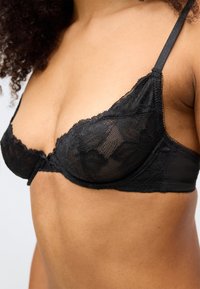 Melnas mežģīņu krūšturis ar caurspīdīgiem ziedu rakstiem, regulējamiem lencēm un aizmugurēju aizdari, izceļot trīsstūrveida krūzītes formu un smalkas detaļas.