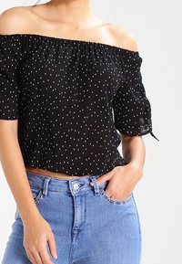 Schwarzes Off-Shoulder-Oberteil mit weißen Polka Dots, kurzen Puffärmeln und gerafftem Halsausschnitt, kombiniert mit hochgeschnittenen blauen Jeans.