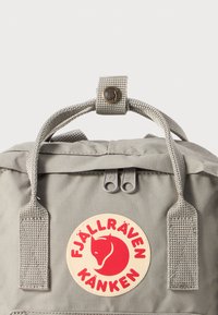 Grå Fjällräven Kånken ryggsäck med justerbara grå webbingremmar, dragkedjereglage och ett framträdande runt logomärke i röd och beige.