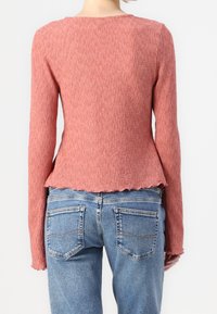 Långärmad korallfärgad stickad topp med en texturerad yta och en vågig nederkant, kombinerad med blå denimjeans med bakfickor.