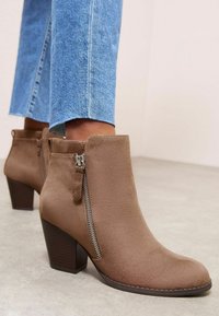 Botas de tobillo de ante marrón con cierre de cremallera y tacón apilado. Diseño acampanado con textura suave y acentos de hardware en contraste.