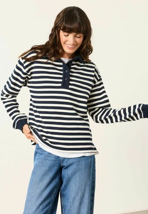 Donna che indossa una camicia a righe blu navy e bianca a maniche lunghe con bottoni, abbinata a jeans blu, sorridente e guardando verso il basso.
