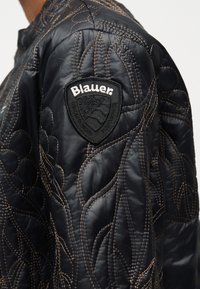 Μανίκι μαύρου καπιτονέ μπουφάν με καφέ ραφές και μαύρο έμβλημα σε σχήμα ασπίδας με την επιγραφή "Blauer" στο πάνω μέρος του βραχίονα.
