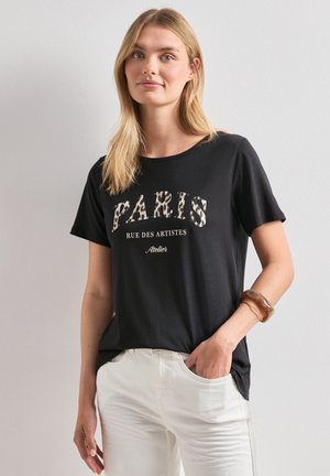 Femme aux cheveux blonds portant un T-shirt noir avec le texte « PARIS RUE DES ARTISTES Atelier » et un pantalon blanc, debout avec une main dans la poche.