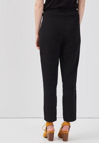 Person wearing black high-waisted tapered pants and mustard yellow heeled sandals standing against a plain light background.