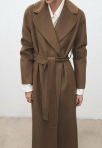 Długi brązowy płaszcz wełniany z paskiem, z klapami z wycięciem i długimi rękawami, noszony na białej koszuli. Gładka faktura i dopasowany krój.