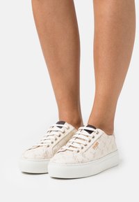 Baskets blanches à plateforme avec un motif étoilé subtil, logo doré sur le côté, portées sur des jambes nues sur un fond clair uni.