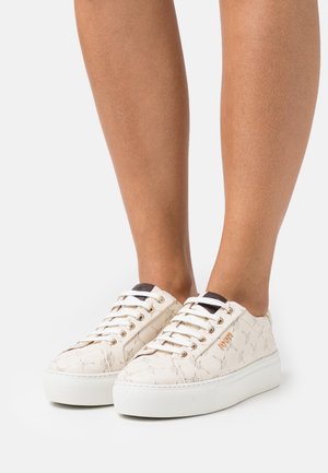 Baskets blanches à plateforme avec un motif étoilé subtil, logo doré sur le côté, portées sur des jambes nues sur un fond clair uni.