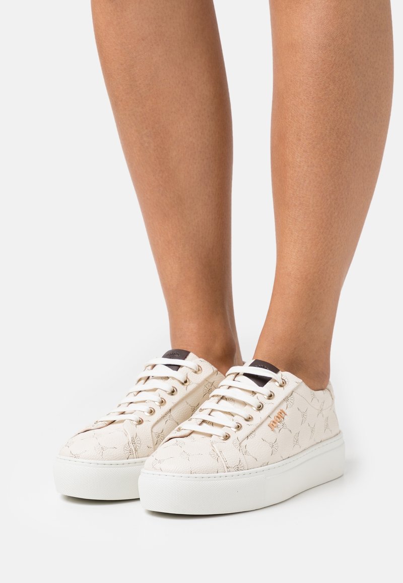 Baskets blanches à plateforme avec un motif étoilé subtil, logo doré sur le côté, portées sur des jambes nues sur un fond clair uni.