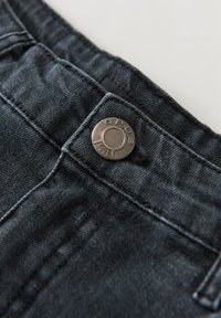 Jeans in denim scuro con un primo piano sulla vita, caratterizzati da un bottone rotondo in argento con la scritta "OPUS" e dettagli di cucitura visibili.