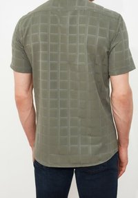 Olivi vihreä kangas, jossa on hienovarainen ruudukkomalli, rento leikkaus ja kaareva helma, näkyy takaa.