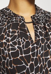 Blusa negra con estampado de jirafa blanco y marrón; presenta un escote plisado y un pequeño acento negro en la abertura. Textura de tela suave.