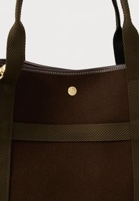 Σκούρο καφέ σουέτ τσάντα με ελαιόχρωμες υφασμάτινες straps και μια μικρή στρογγυλή χρυσή λεπτομέρεια κοντά στην επάνω άκρη.