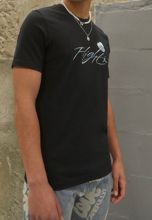 Personne portant un t-shirt noir avec un graphisme "Flight", un jean bleu clair avec un texte blanc, et des colliers en argent et perles superposés devant un mur en pierre.