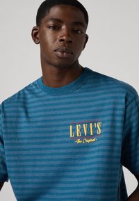 T-shirt azul riscado com neckline redonda e um logotipo frontal em vermelho e amarelo com o texto "LEVI'S The Original." Textura suave.
