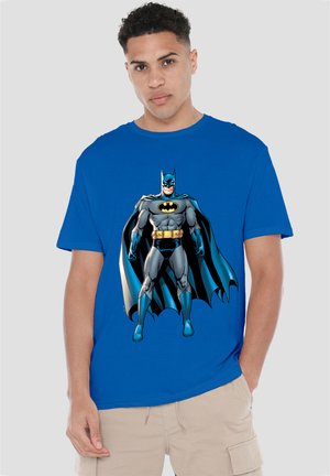 Young man wearing a blue t-shirt with a detailed Batman graphic and beige cargo shorts standing against a plain background.