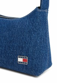 Denim schoudertas met een driehoekige vorm, donkerblauwe kleur, witte stiksels en een metalen logo plaat met "TOMMY JEANS" in vette letters.