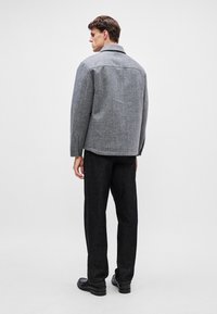 Grau melierte Wollmischjacke mit Kragen, lockerer Passform und Rückenpasse, kombiniert mit dunklen Hosen und schwarzen Schuhen. Minimalistisches Design.