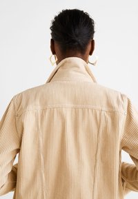 Beige corduroy jack met verticale ribbels, spitse kraag en een casual pasvorm. Heeft subtiele stiksels en drukknoopsluitingen.