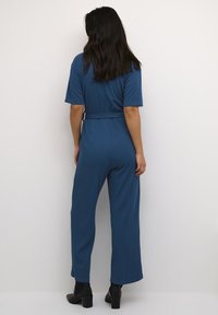 Mono de canalé azul con cintura ceñida, mangas cortas y diseño de pierna ancha. Botas negras hasta el tobillo completan el atuendo.