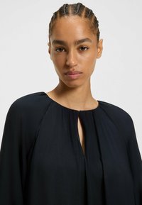 Svart plagg med en lös passform, rund halsringning och en nyckelhålsdetalj framtill. Tillverkat av ett strukturerat tyg med subtila veck.