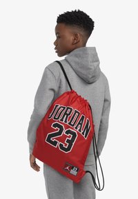 Rød trekkveske med "JORDAN" og "23" trykket i svart. Laget av glatt stoff med en blank overflate og svarte stropper.