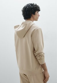 Sweat à capuche beige en tissu doux, avec une coupe décontractée, des manches longues et une capuche avec cordon de serrage. Design simple avec des poignets et un ourlet côtelés.