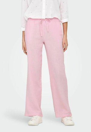 Jasnoróżowe szerokie spodnie z teksturowanej tkaniny, z regulowanym paskiem na sznurki w pasie i luźnym krojem. Noszone z białymi trampkami.