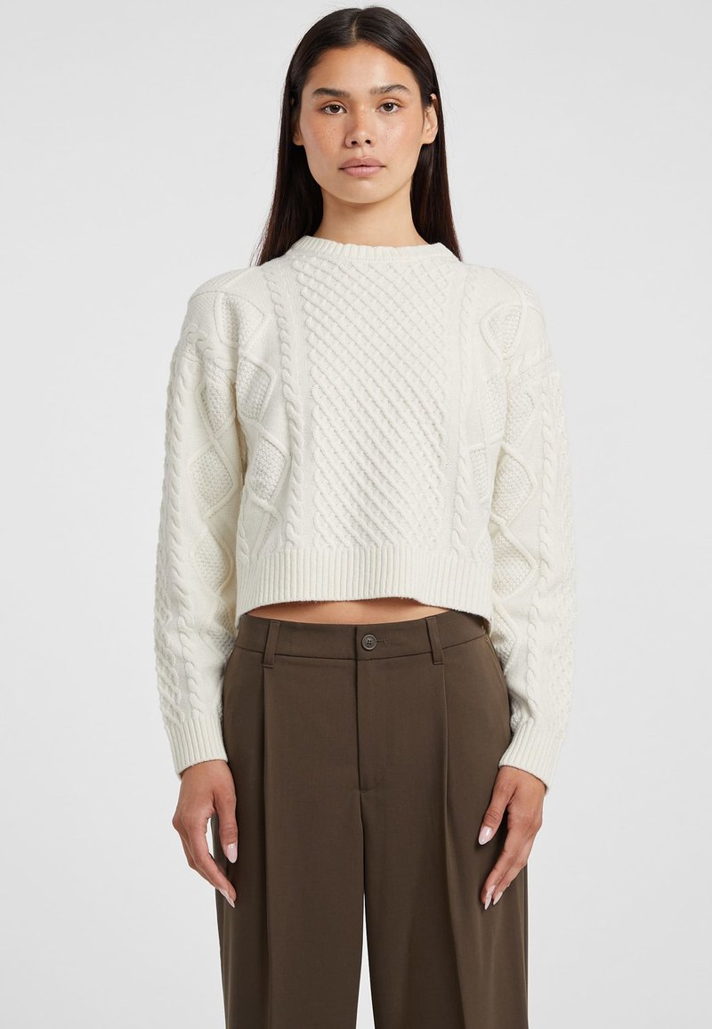 Maglione corto color crema con design a trecce testurizzato e polsini a coste. Abbinato a pantaloni tailored marroni a vita alta.