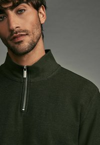 Tmavozelený pulóver na zips z textúrovaného materiálu. Má vysoký golier a strieborný zips na krku.
