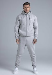 Gray zip-up hoodie with drawstrings and small logo, paired with matching joggers in a soft texture, worn with white sneakers.