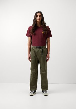 Camiseta marrón con pequeño logo amarillo, pantalones verde oliva con cintura elástica y bolsillo lateral, combinados con zapatillas negras.