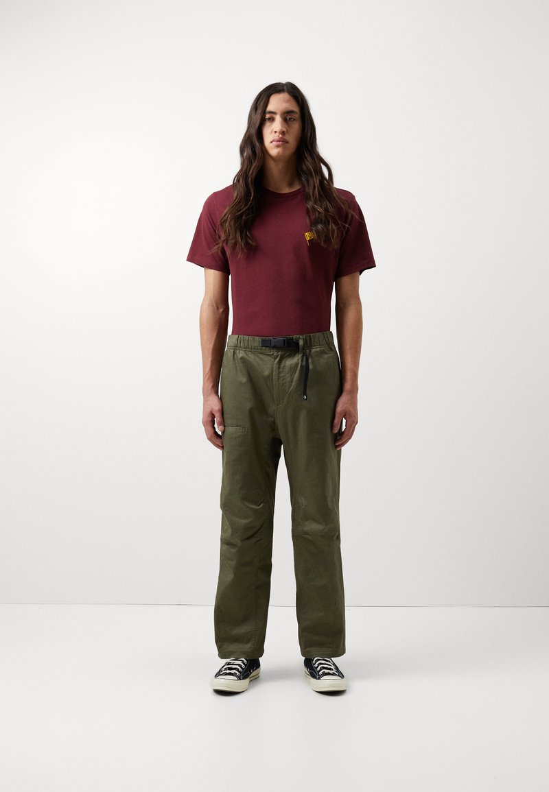 Camiseta marrón con pequeño logo amarillo, pantalones verde oliva con cintura elástica y bolsillo lateral, combinados con zapatillas negras.