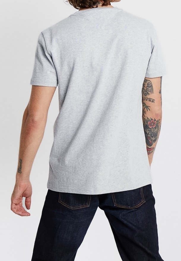 Homme portant un t-shirt à manches courtes gris clair uni et un jean bleu foncé, montrant son dos avec des tatouages sur l'avant-bras droit et le bras supérieur.