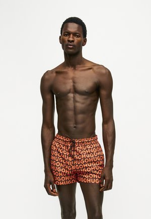 Homme mince à la peau foncée debout, torse nu, portant un short orange et noir avec un motif de texte répétitif "HUGO" sur fond blanc.