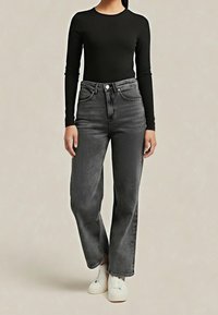 Mujer con camiseta negra ajustada de manga larga, jeans grises de cintura alta y zapatillas blancas, de pie contra un fondo liso.