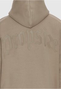 Sweat à capuche beige avec texte en relief "Dropsied" en police de style gothique sur le haut du dos.