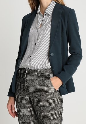 Femme portant un blazer bleu marine sur une chemise blanche et noire à rayures avec un pantalon à carreaux noir et blanc, main dans la poche.