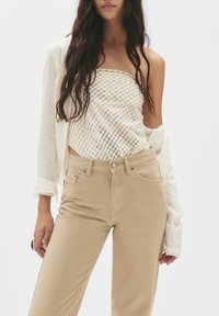 Crème-kleurige tube top met een ruitpatroon, gecombineerd met beige broek met hoge taille. Een lichte, gestructureerde oversized jas is over de schouder gedrapeerd.