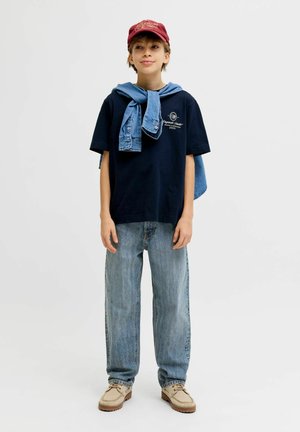 Junge Person steht mit entspannten Händen, trägt rote Mütze, schwarzes T-Shirt, hellblaue Jeans, beige Schuhe und eine Jeansjacke, die über die Schultern gelegt ist.