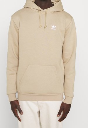 Personne portant un sweat à capuche beige Adidas avec poche avant et cordon de serrage, associé à un pantalon écru sur fond blanc.