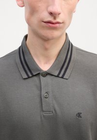 Šedé polo tričko s texturovanou tkaninou, dvěma knoflíky a límcem se tmavě šedými pruhy. Malé logo na hrudi.