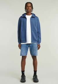 Un sweat à capuche zippé bleu en matière douce avec un cordon de serrage, associé à un short en denim clair et des chaussures de sport noires. Deux poches avant.